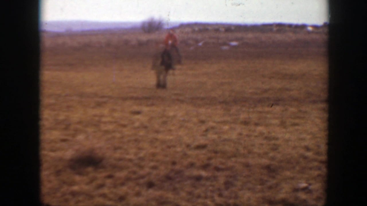 A child in a bright red jacket appears to ride a horse toward the camera across a broad Montana field.