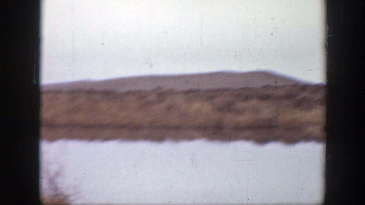 A brief Montana landscape cutaway shows calm water, a grassy bank, and low hills beneath a pale sky.