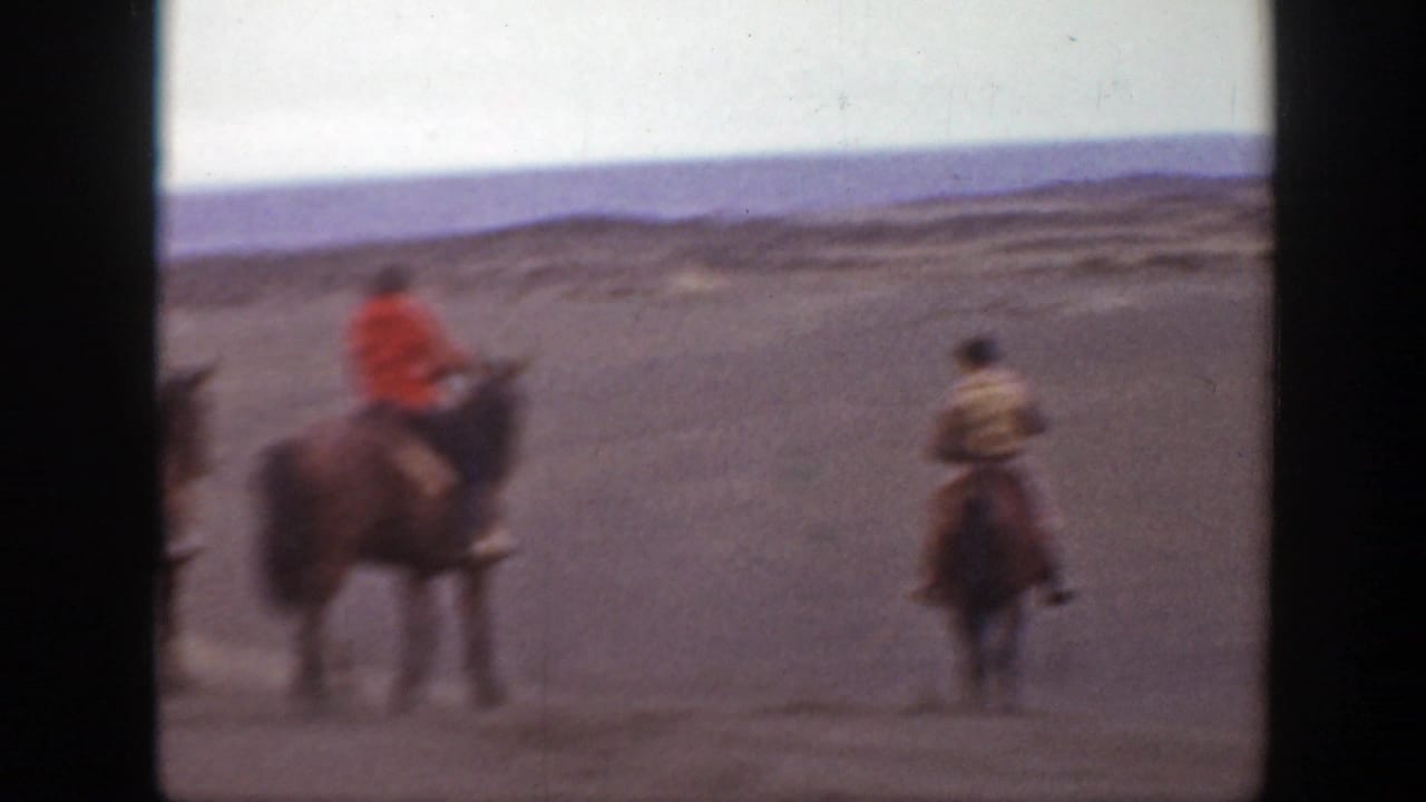 Three riders on horses cross a flat open landscape, with one rider in a red jacket.