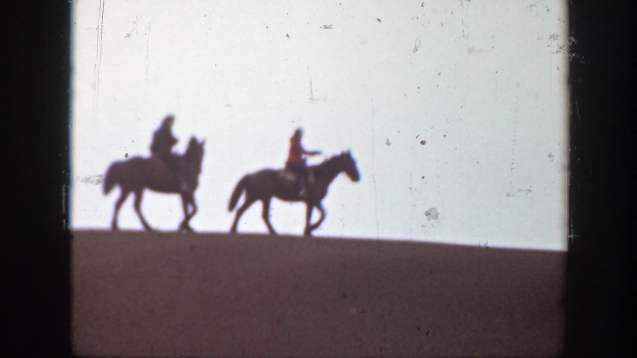 Two riders on horseback continue across the ridge in silhouette.
