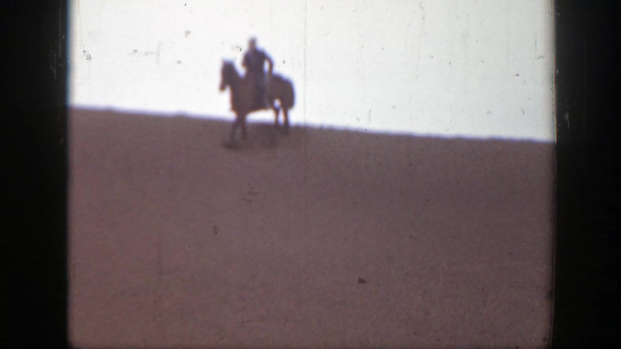 Mounted rider pauses near the top of a sandy slope with another horse partly visible nearby.