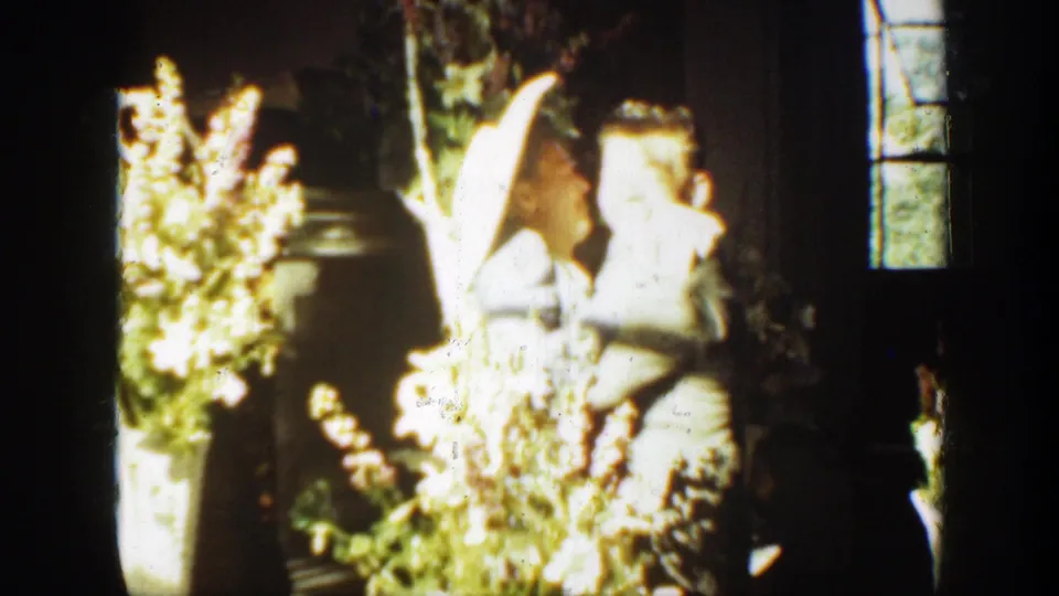 An overexposed 8mm frame appears to show a person near flowers inside or beside a darkened space, with a window visible at the right edge.