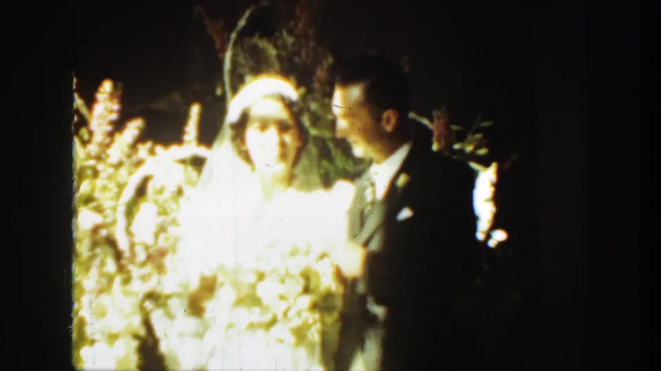 1939 Montana home-movie frame of a bride and groom standing close in strong light, with flowers and dark foliage behind them.