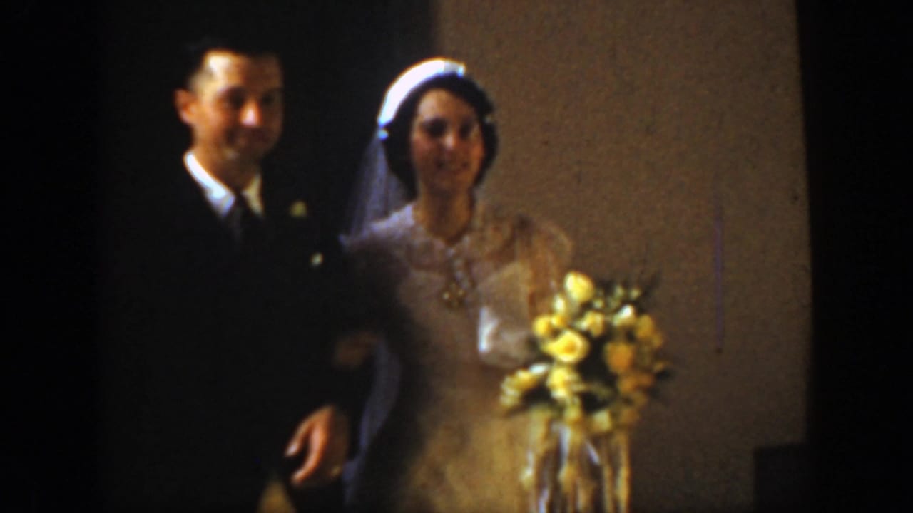 In this 1939 Montana home movie frame, a bride in a veil stands beside a groom in a dark suit, holding a yellow bouquet indoors.