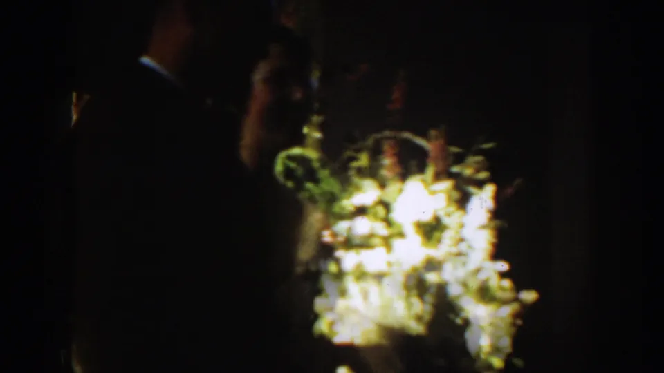 A dark, grainy 1939 Montana wedding frame shows floral displays and shadowed guests during the indoor ceremony.