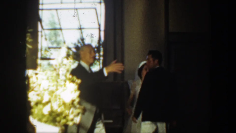 In a 1939 Montana wedding interior, a man in formalwear gestures near a large window while the bride and groom stand to the side.