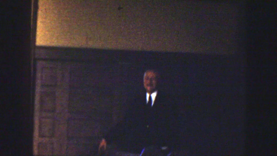Adult man in a dark suit appears in a dim 1939 Montana wedding interior, partly obscured by low exposure.