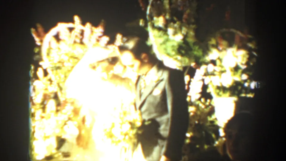 Bride in white and groom in a dark suit stand among bright wedding flowers in 1939 Montana home-movie footage.