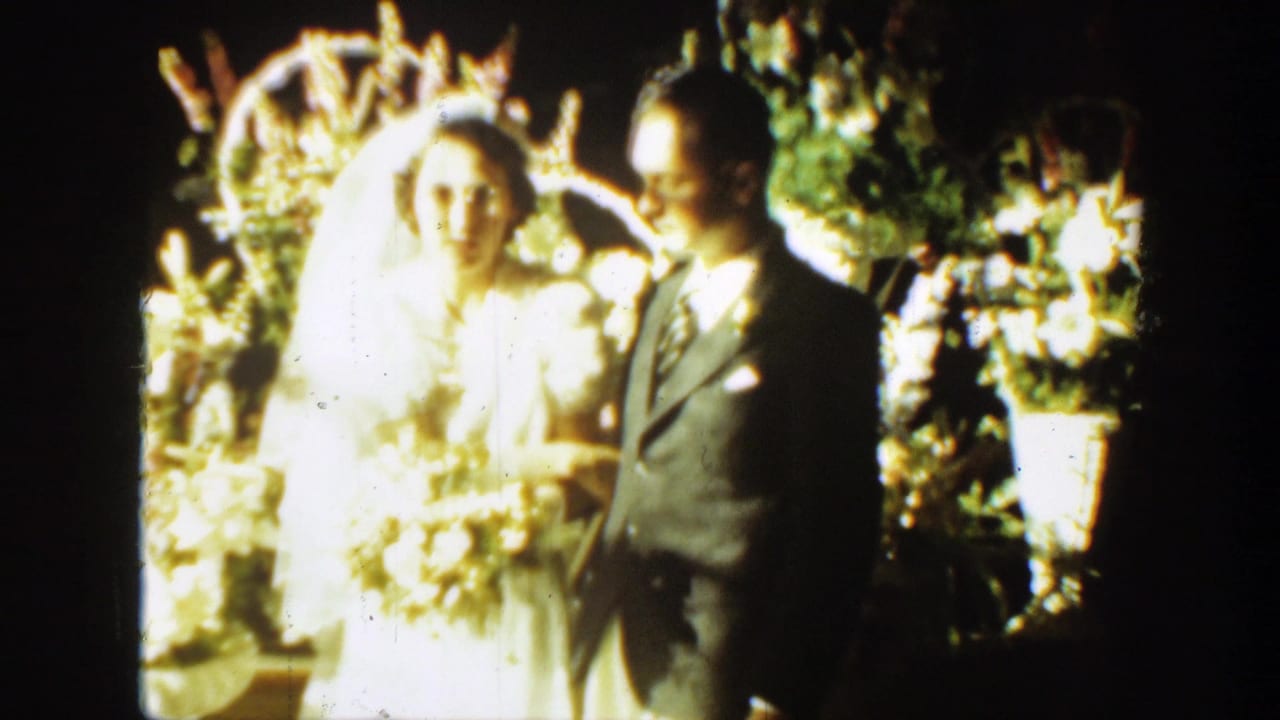 In 1939 Montana footage, a bride in a white dress and veil stands with a bouquet beside a groom in a dark suit near a floral arch.
