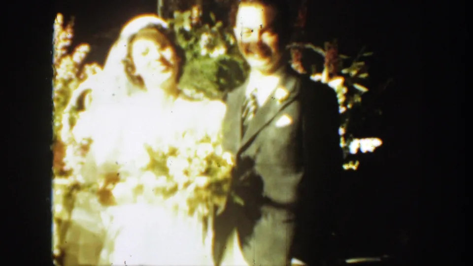 1939 Montana home-movie frame of a bride in a veil beside a groom in a suit under a floral arch.
