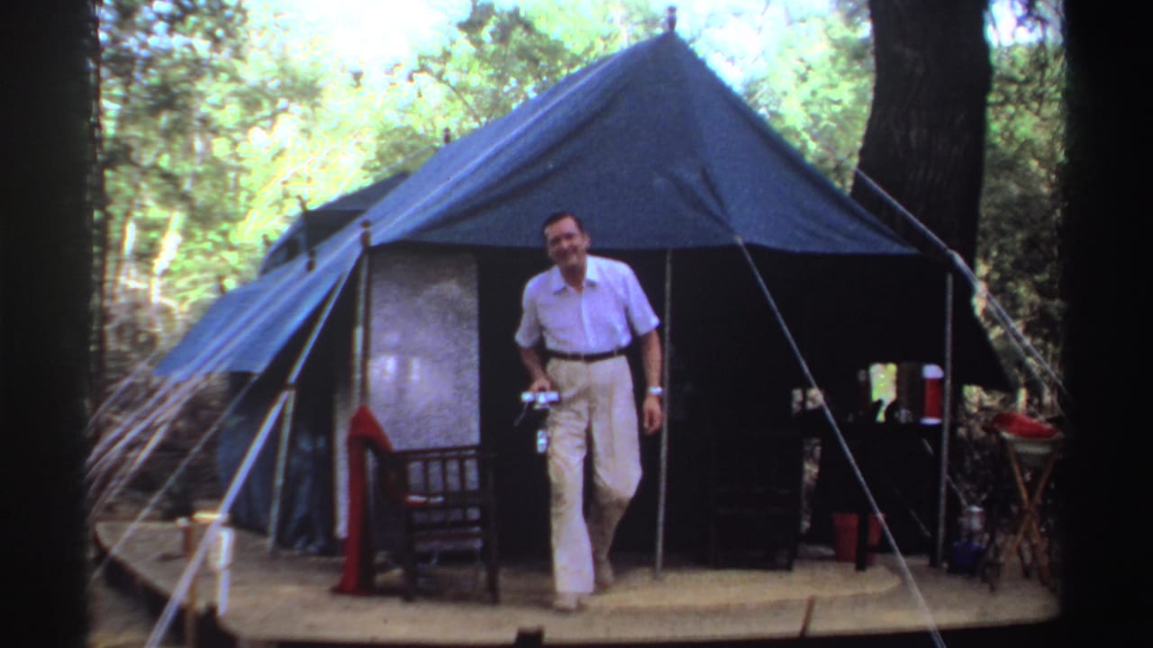 1969 Sudan 8mm frame showing an adult man emerging from a blue canvas tent while holding a camera near trees.