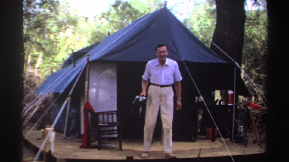 1969 Sudan 8mm frame showing an adult man standing before a blue camp tent, holding a camera on a wooded platform.