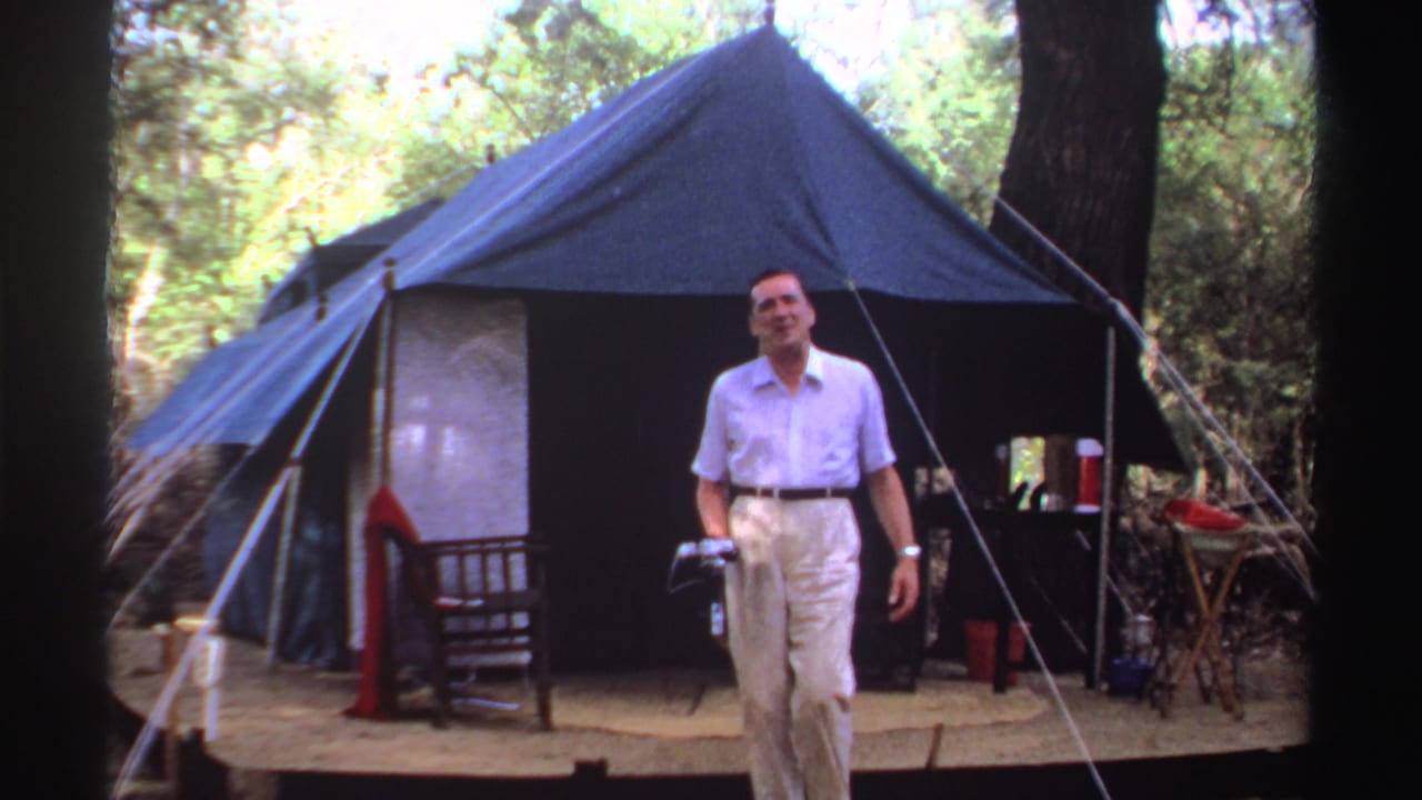 Man in light clothing stands outside a blue canvas tent in a wooded area.