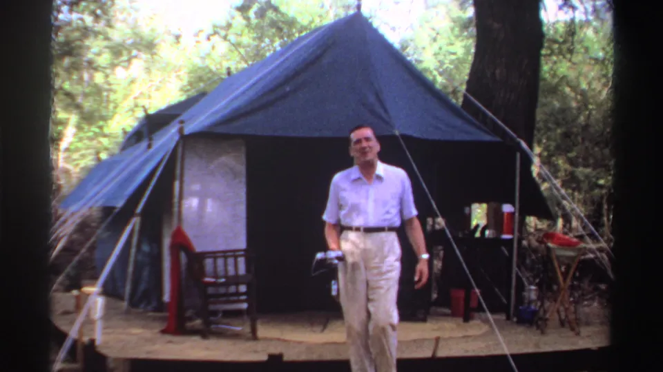 In Sudan 1969 home-movie footage, a man stands smiling near a blue canvas tent with trees, ropes, and camp items around him.