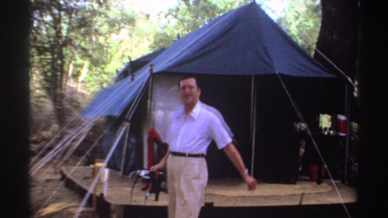 Man walks toward the camera from a canvas tent while holding a small camera.