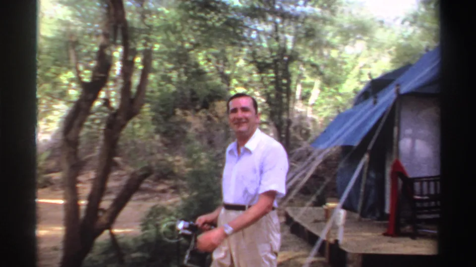 The man smiles while holding a camera near trees and the side of a blue canvas tent in the 1969 Sudan camp scene.