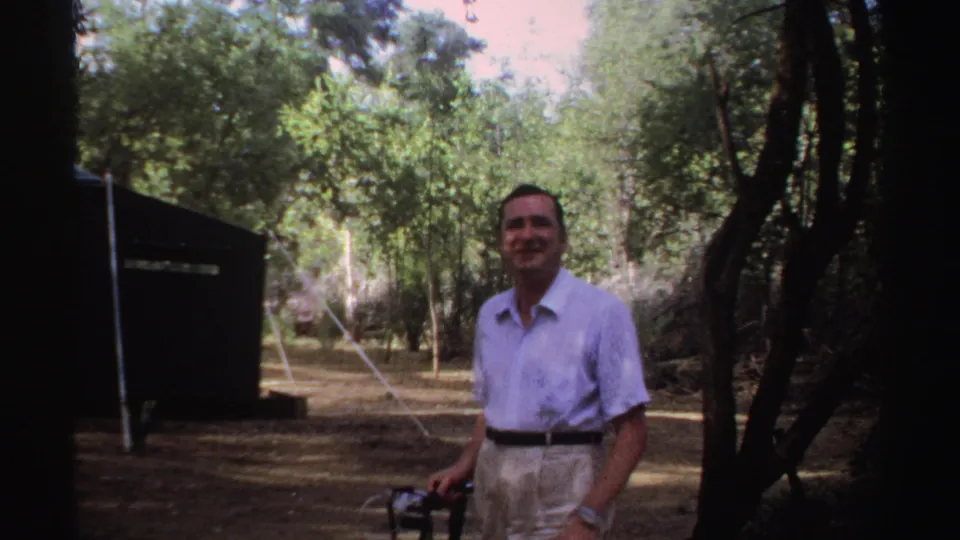 A smiling man holding a camera stands among trees, with the dark side of the tent and ropes behind him in Sudan 1969 footage.