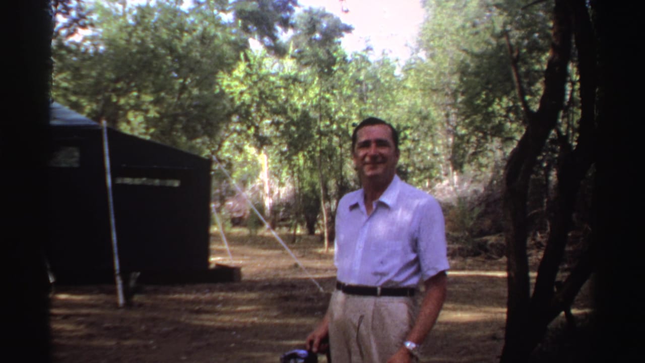 Man in a light shirt stands smiling among trees beside a dark tent.