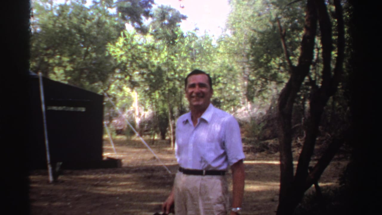 The man remains facing the camera in the Sudan campsite, with trees, dry ground, and a dark tent visible behind him.