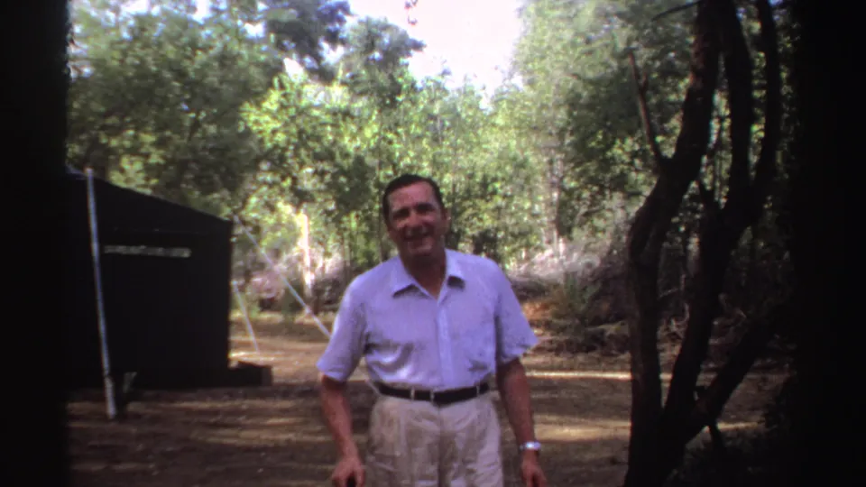 The man shifts his posture in the wooded Sudan campsite, with the tent at left and dense trees behind him.