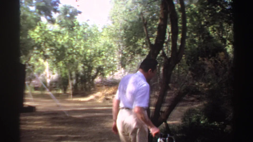 The man turns from the camera and walks toward a tree in the Sudan campsite, holding a small camera at his side.