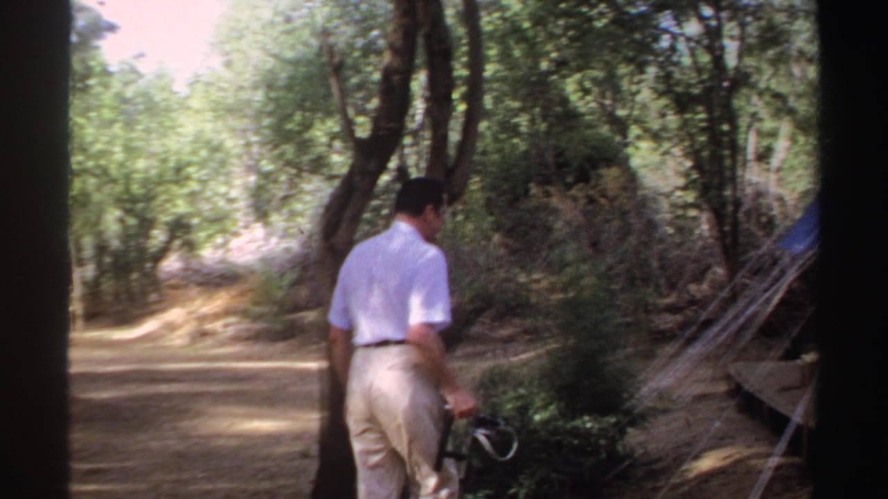 Man in white shirt walking on a wooded path beside a tent.