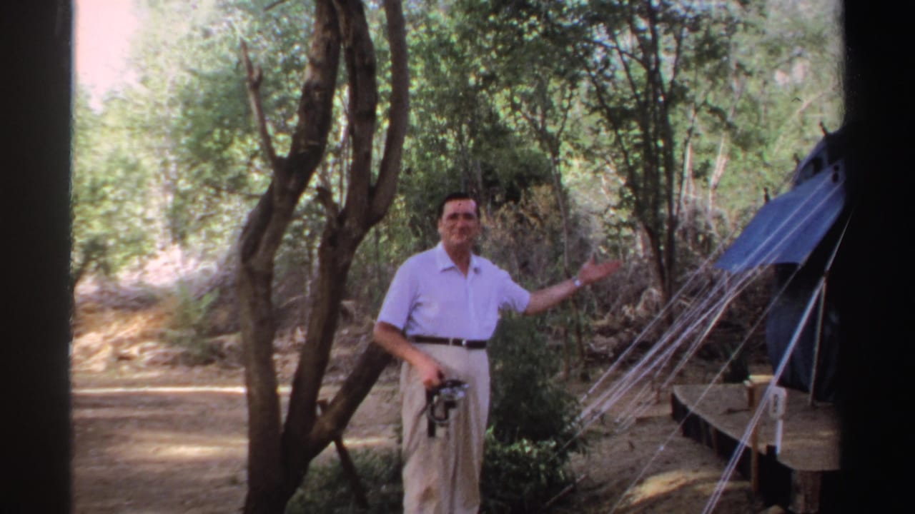 In the wooded Sudan campsite, the man faces the camera and gestures toward a blue tent under trees.