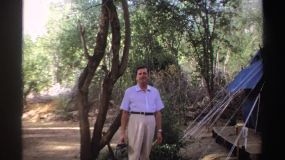 The 1969 Sudan home-movie frame shows the man standing front-facing near a blue tent and trees, with a camera in one hand.