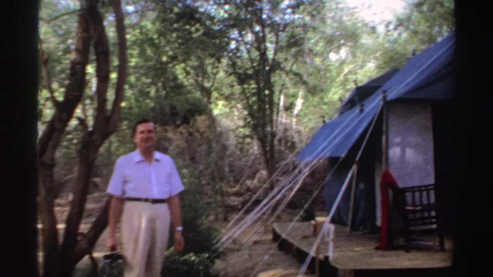 The man remains near the blue tent in the wooded Sudan campsite, framed by trees and 8mm film borders.