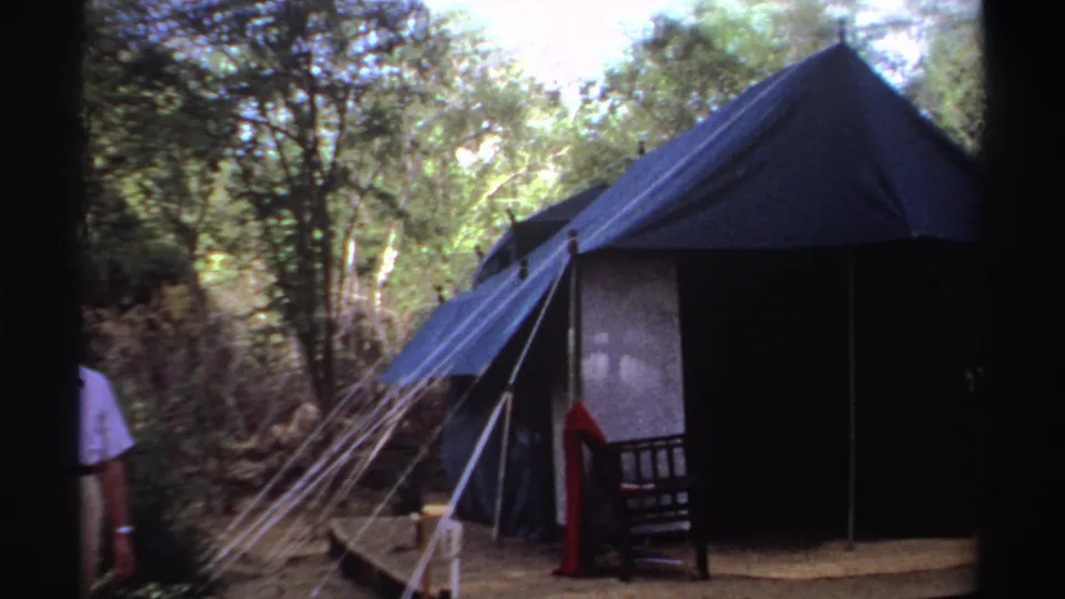 1969 Sudan 8mm frame showing a man at the edge of a forest campsite with blue canvas tents, ropes, and a wooden platform.