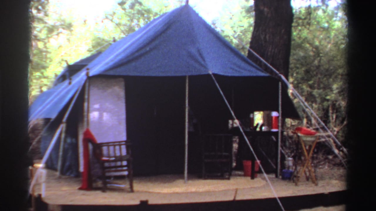 1969 Sudan campsite frame with a blue canvas tent, porch chair, table with red containers, ropes, and surrounding woodland.