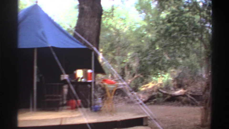1969 Sudan 8mm frame showing the edge of a blue canvas tent on a platform with a chair, camp table, ropes, and dense brush nearby.