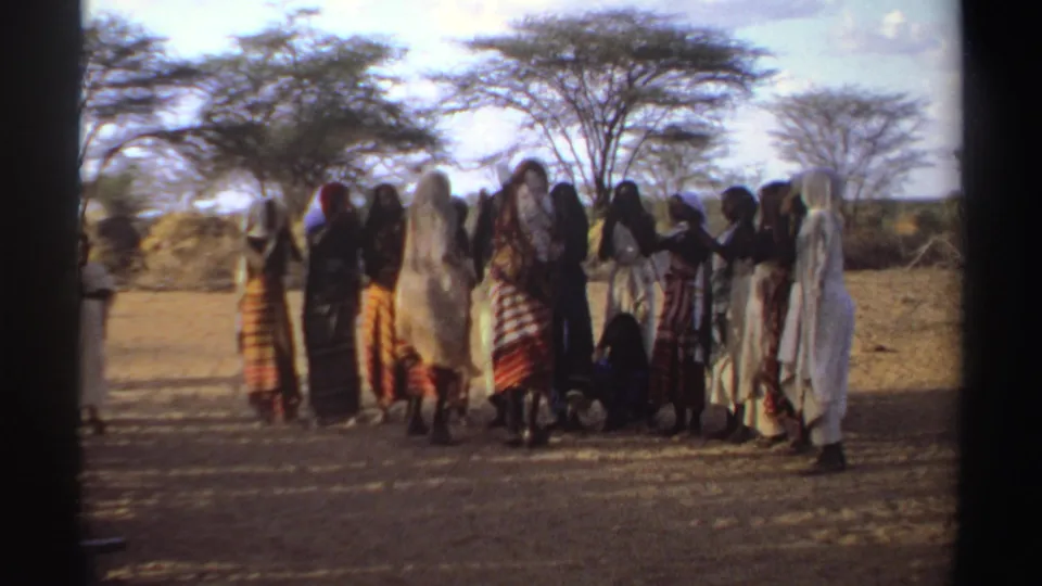 1969 Sudan home-movie frame of women in patterned dresses and white wraps gathered outdoors beneath sparse trees.