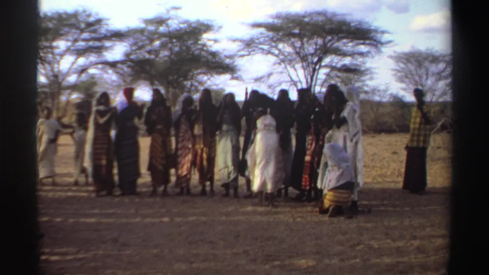 1969 Sudan frame of women gathered in a communal dance or clapping circle, seen on faded 8mm film beneath sparse trees.