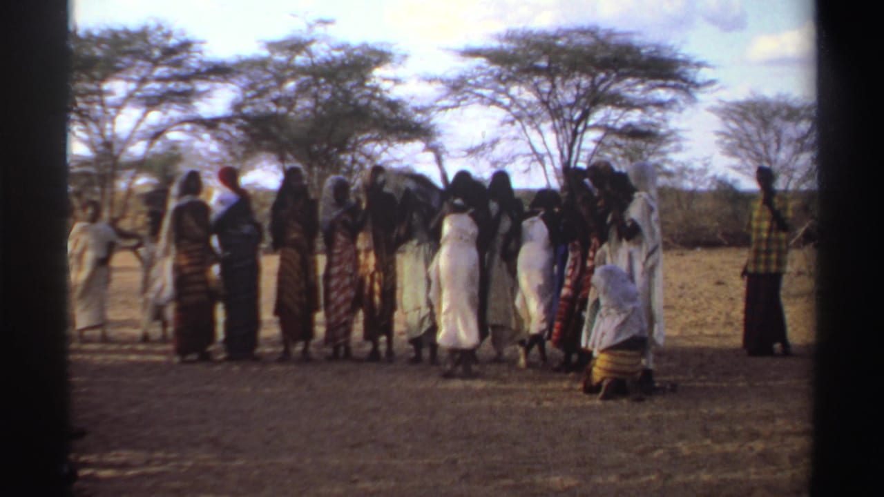 1969 Sudan 8mm frame showing women in traditional dresses gathered in a dry landscape with trees behind them.
