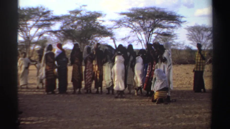 1969 Sudan 8mm frame showing women in traditional dresses gathered in a dry landscape with trees behind them.