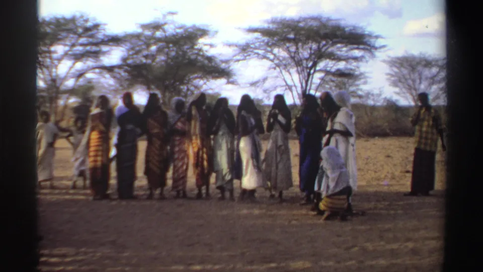 Women in Sudan, 1969, stand in a loose line outdoors, with headscarves, patterned dresses, and trees in the background.