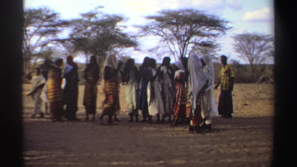 A group of women stands in an arid Sudanese field while a blurred figure passes across the 8mm frame.