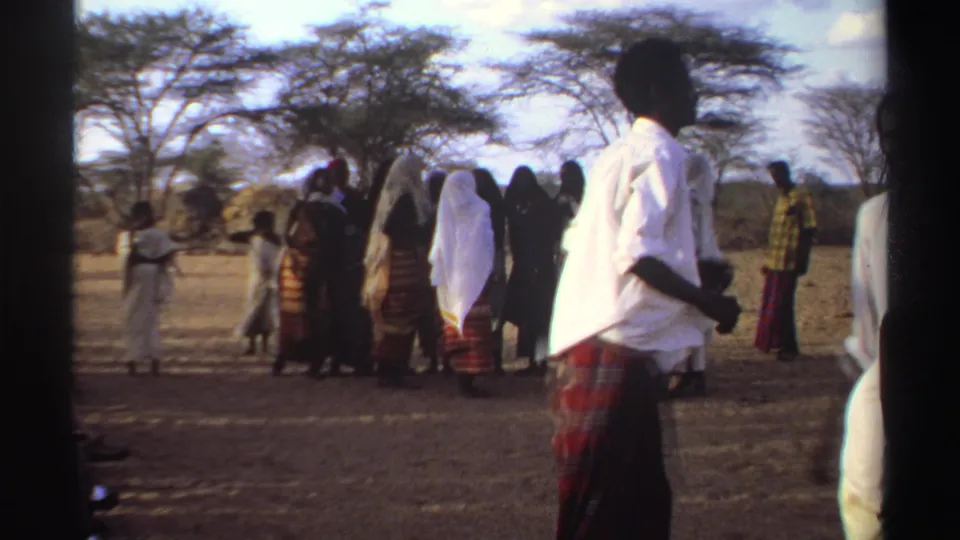 A man in a white shirt appears in the foreground as women stand together in a dry Sudanese field in 1969 home-movie footage.