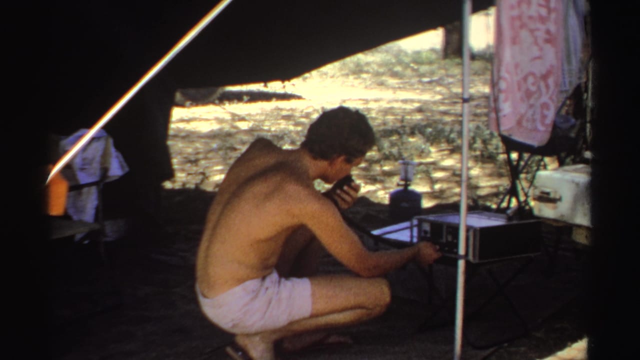 Shirtless man crouching under a tent beside portable radio equipment.