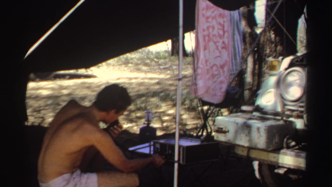 A shirtless man in Sudan in 1969 appears to adjust radio equipment beside a parked car under a shaded camp cover.