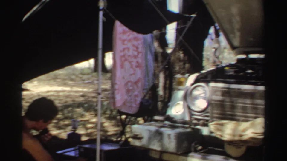 The frame shows the man partly cropped at left, radio equipment in shade, and a car front with its hood raised in Sudan in 1969.