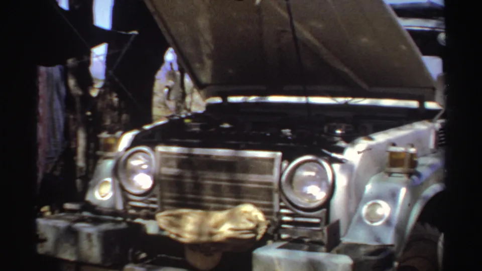 A vintage car stands under shade with its hood open, camp items and fabric visible nearby in the 1969 Sudan home movie.