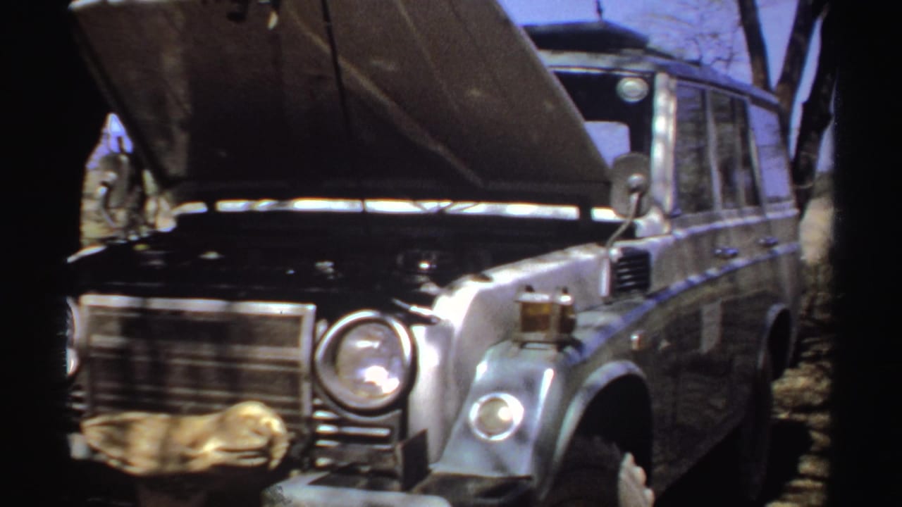 Silver utility vehicle with its hood raised on dry ground near trees.