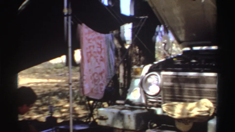 1969 Sudan camp frame showing the vehicle front beside canvas shade, poles, containers, and a patterned hanging cloth.