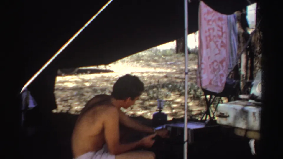 1969 Sudan home-movie frame showing a shirtless man seated under canvas shade beside camp supplies and a hanging cloth.