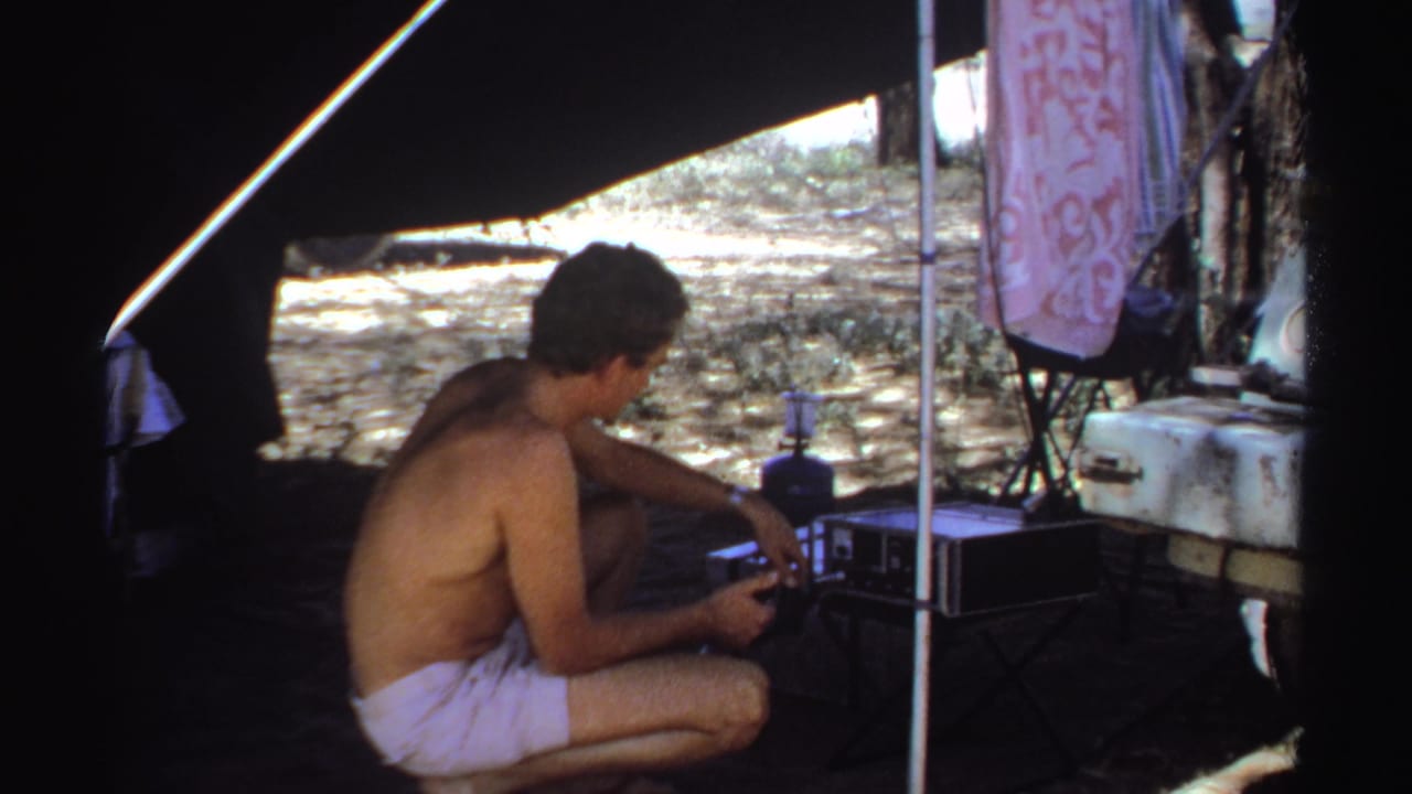 Shirtless man crouching under a tent beside camp equipment and a patterned hanging cloth.