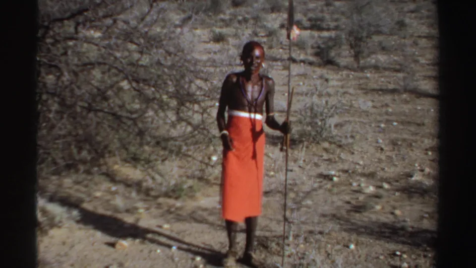 1969 Sudan 8mm frame of a man in orange shuka-style cloth holding a long spear in dry scrubland.