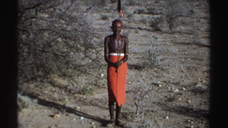 1969 Sudan 8mm frame of the man centered in dry scrubland, holding a spear and facing the camera.