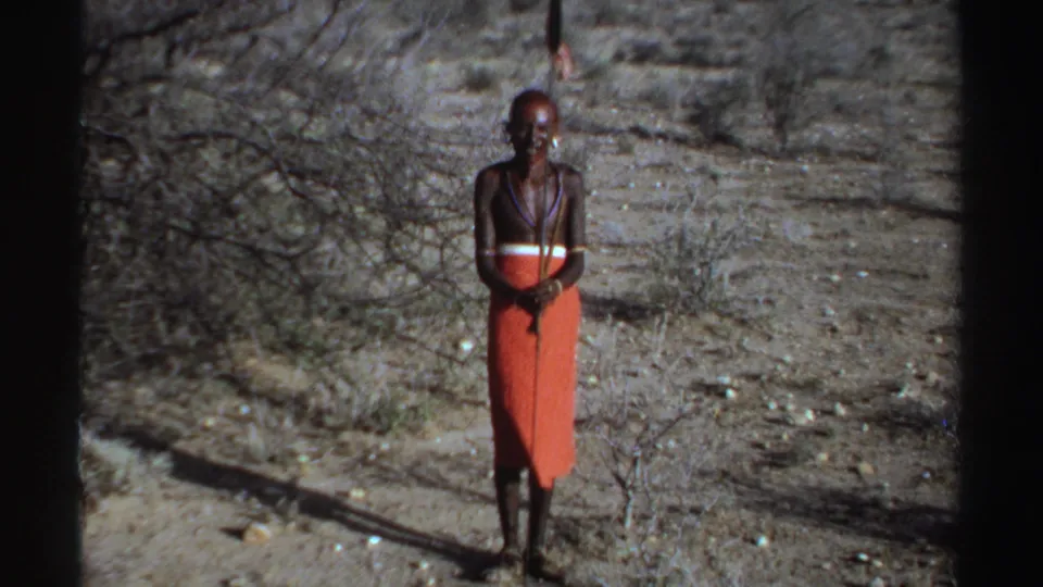 1969 Sudan-labeled 8mm frame showing a lone adult man in red wrapped cloth, bead necklaces, and a spear in an arid landscape.